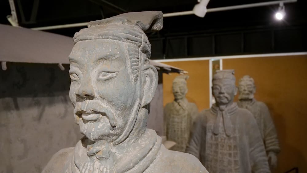 Close-up of a life-sized terracotta warrior replica's face and headdress in the Chinese Artifacts Museum in Calgary, Alberta, Canada, with several more warrior figures visible in soft focus against an orange gallery wall.