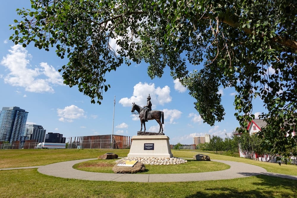 Estatua ecuestre y parque de The Confluence y antiguo Fort Calgary en Calgary, Alberta, Canadá Estatua ecuestre en los espacios verdes de The Confluence, Calgary, Alberta, Canadá, enmarcada por árboles frondosos, con edificios modernos de la ciudad y el sitio histórico al fondo.