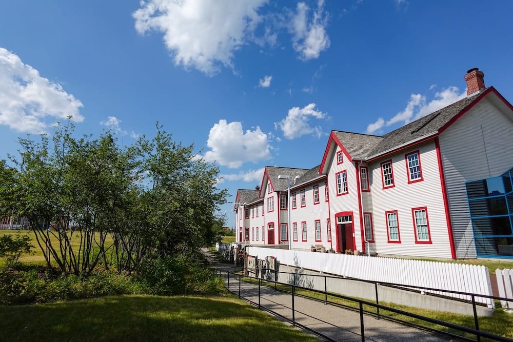 Edificios históricos y terrenos de The Confluence en Calgary, Alberta, Canadá Edificios históricos blancos con detalles rojos en The Confluence, Calgary, Alberta, Canadá, junto a un sendero pavimentado accesible, jardines verdes y árboles bajo un cielo azul de verano.