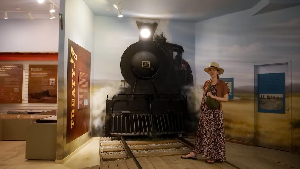 Audrey Bergner en una exhibición ferroviaria de The Confluence en Calgary, Alberta, Canadá Audrey Bergner junto a una locomotora de vapor dentro de The Confluence, Calgary, Alberta, Canadá, con vías, paneles interpretativos y material sobre Treaty 7 alrededor.