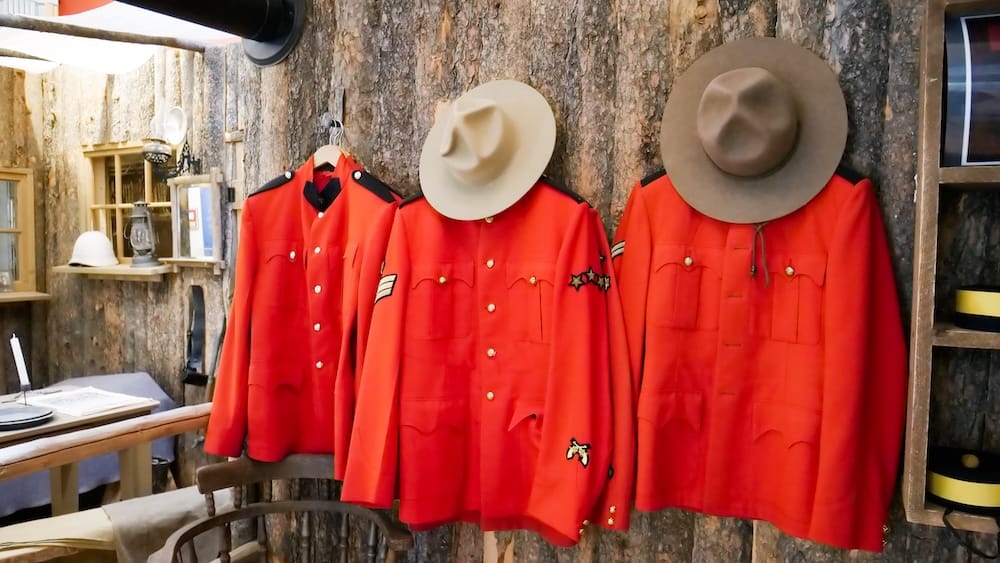 Three red RCMP-style uniforms and wide-brimmed hats hang against a rustic wood wall inside The Confluence historic site in Calgary, Alberta, Canada, with period furnishings and display shelves nearby.