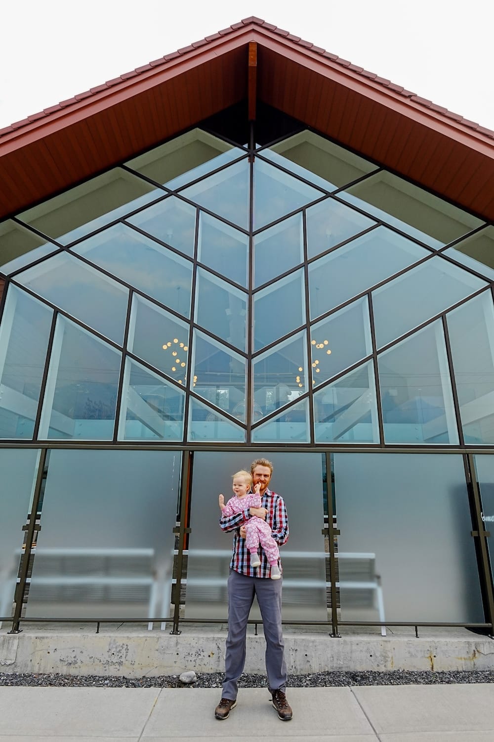 Samuel Jeffery holds Aurelia Jeffery outside The Gem in Harvie Heights, Alberta, Canada, in front of the accommodation’s large glass facade and steep peaked roof.