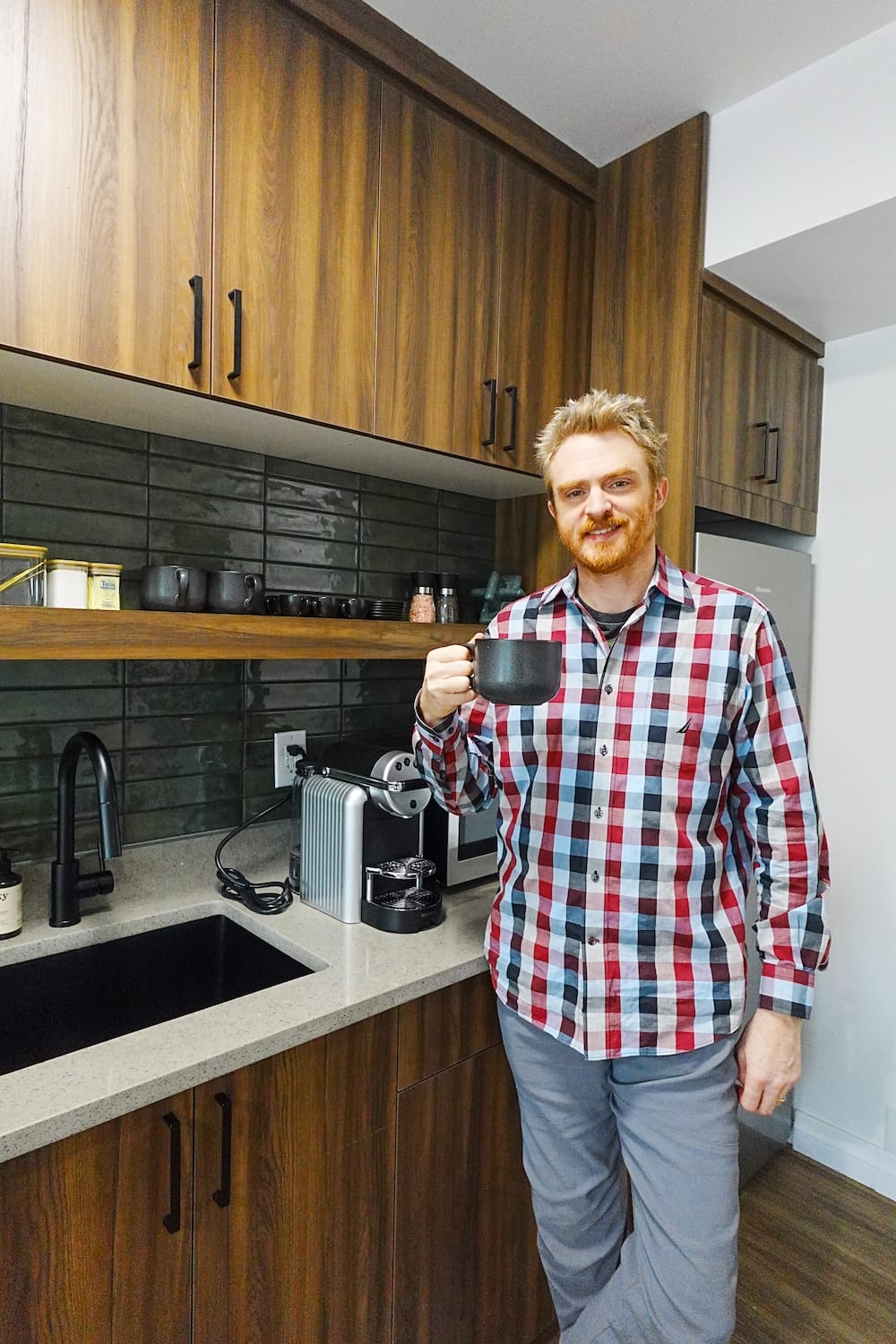 Samuel Jeffery holds a mug beside the kitchenette at The Gem in Harvie Heights, Alberta, Canada, with wood cabinetry, a sink, coffee machine, cups and refrigerator visible behind him.