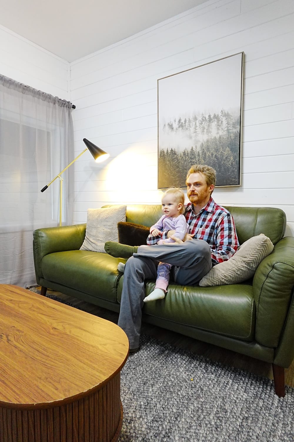 Samuel Jeffery sits with Aurelia Jeffery on a green sofa in the living room at The Gem in Harvie Heights, Alberta, Canada, with a floor lamp, coffee table and framed forest artwork showing the family-friendly suite interior.