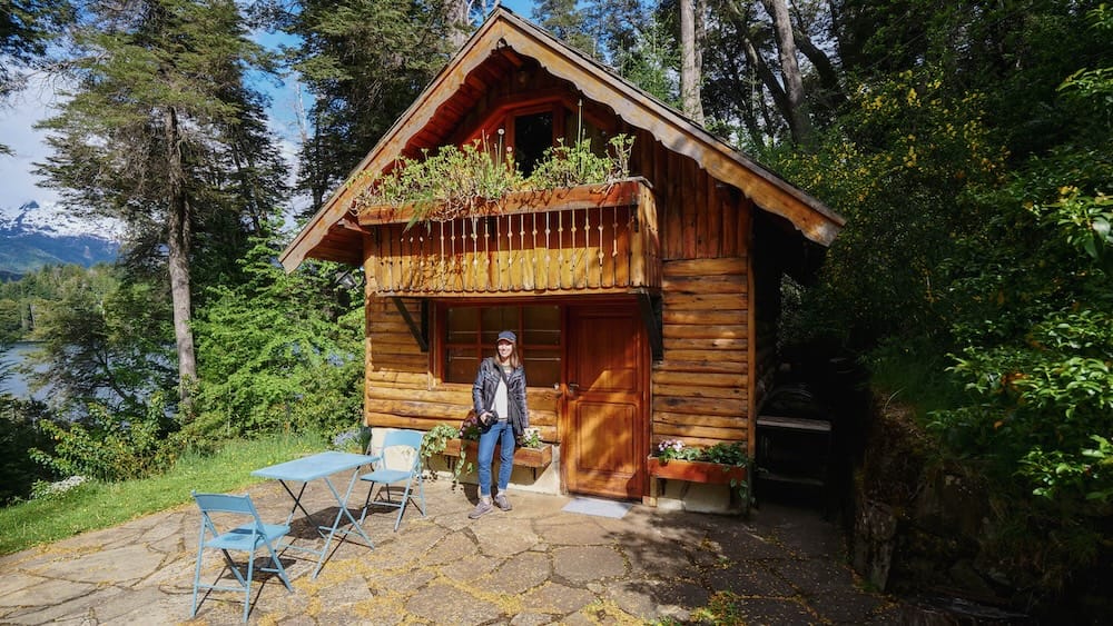 Audrey Bergner stands outside a timber cabin near Bariloche, Río Negro, Patagonia, Argentina, with a wooden balcony, patio furniture, dense forest, a blue lake and snow-dusted mountains visible behind her.