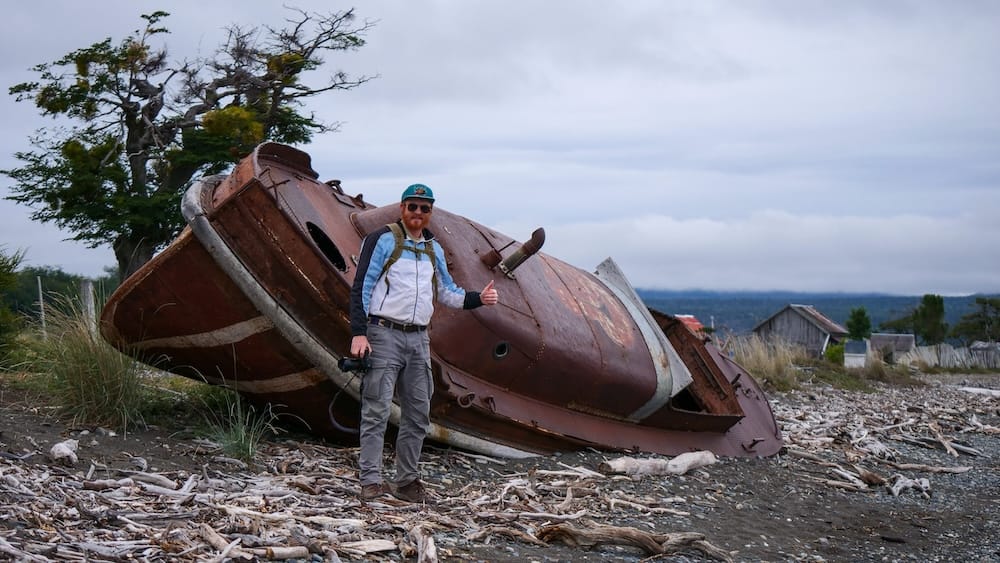 Tolhuin Tierra del Fuego Shipwreck and Patagonia Weather Season Choice with Samuel Jeffery Samuel Jeffery beside a weathered shipwreck on the coast near Tolhuin, Tierra del Fuego, showing how Patagonia’s seasonal mood can shift toward cold wind, grey skies and rugged coastal exploring far beyond the classic summer brochure.