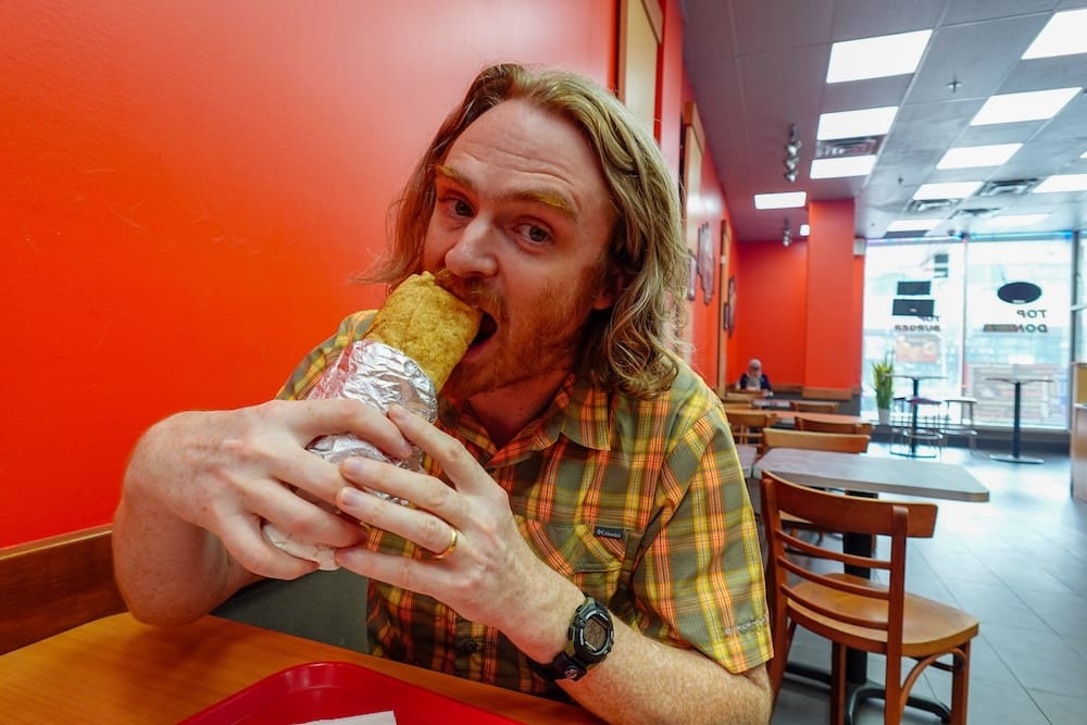 Samuel Jeffery Eating a Donair at Top Donair in Edmonton, Alberta, Canada Samuel Jeffery bites into a large foil-wrapped donair while seated inside Top Donair in Edmonton, Alberta, Canada, with orange walls, wooden tables and the restaurant entrance behind him.