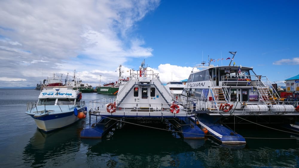 Embarcaciones de fauna en el puerto de Ushuaia, Tierra del Fuego, Patagonia, Argentina, antes de salir al Canal Beagle Varias embarcaciones de excursión están amarradas en el puerto de Ushuaia, Tierra del Fuego, Patagonia, Argentina, con catamaranes, equipos portuarios y barcos mayores bajo un amplio cielo azul con nubes.