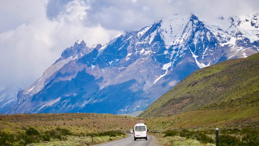 Combi de tour acercándose al macizo de Torres del Paine, Región de Magallanes, Chile Una combi blanca de tour avanza por un camino de ripio hacia el macizo escarpado y parcialmente nevado de los Cuernos del Paine bajo un cielo muy nublado, con matorral de estepa verde en primer plano, Parque Nacional Torres del Paine, Región de Magallanes, Patagonia chilena.