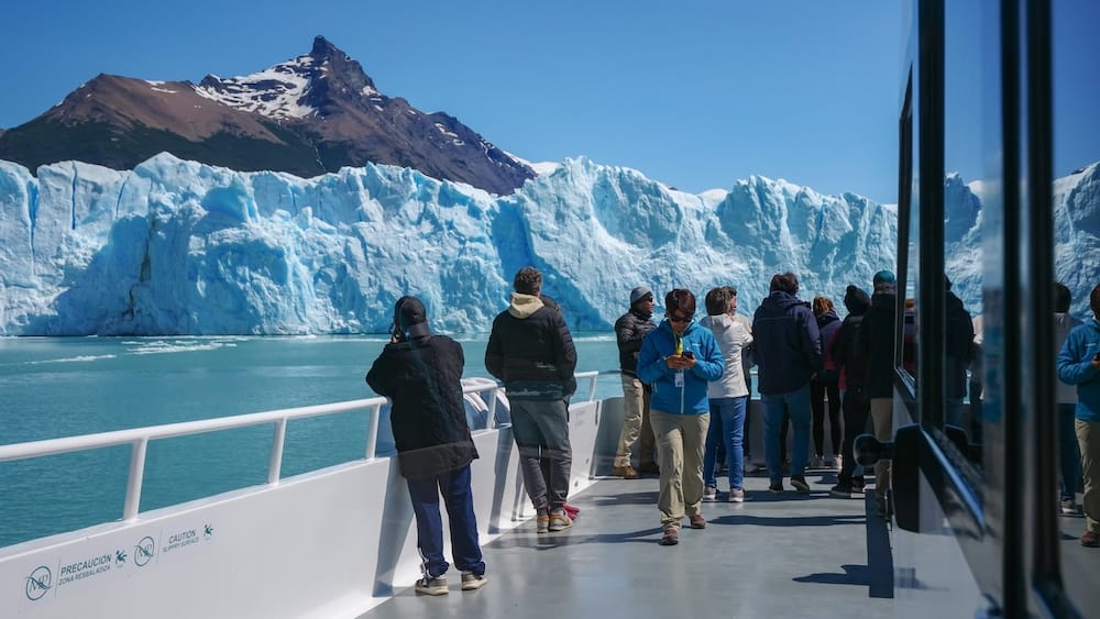 Tourists aboard a boat beside Perito Moreno Glacier in Santa Cruz, Patagonia, Argentina Tourists stand on the outdoor deck of a glacier boat near Perito Moreno Glacier in Santa Cruz, Patagonia, Argentina, with the towering blue ice wall and a rugged mountain peak behind them.