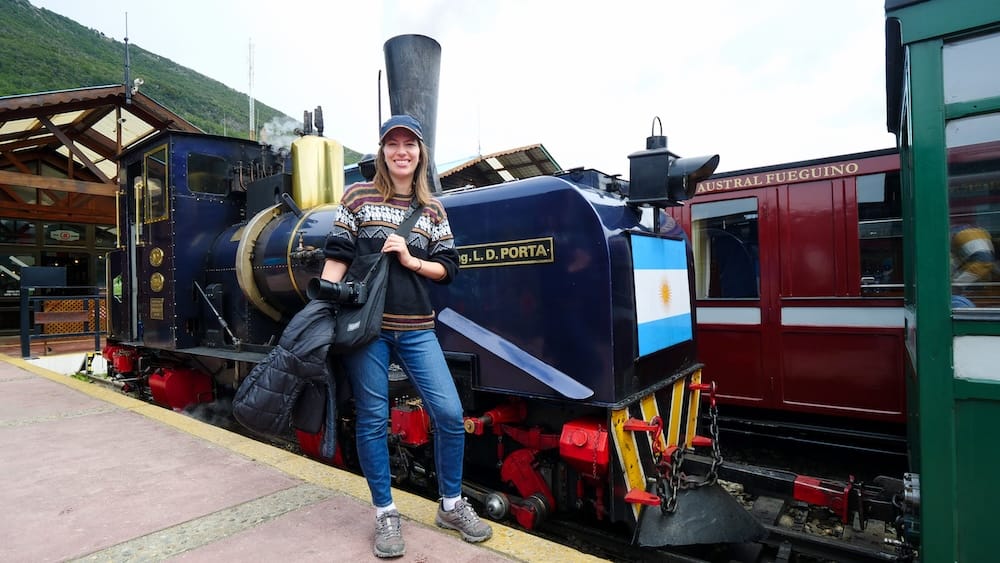Audrey Bergner stands beside a historic steam locomotive at the End of the World Train station near Ushuaia, Tierra del Fuego, Patagonia, Argentina, with colourful carriages and forested hills behind her.