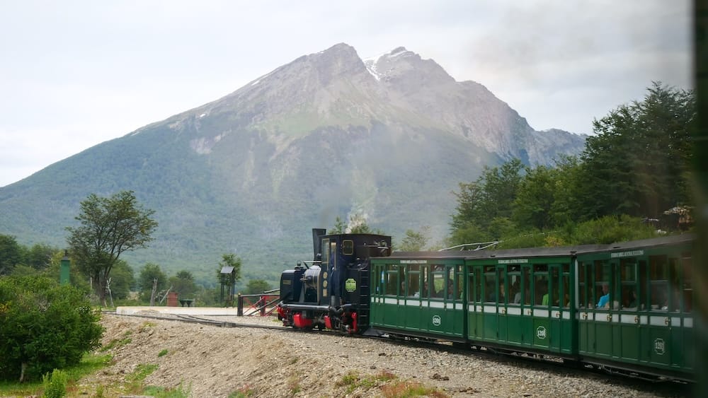 The Train at the End of the World travels beneath a forested mountain near Ushuaia, Tierra del Fuego, Patagonia, Argentina, with a black steam locomotive pulling green heritage carriages.