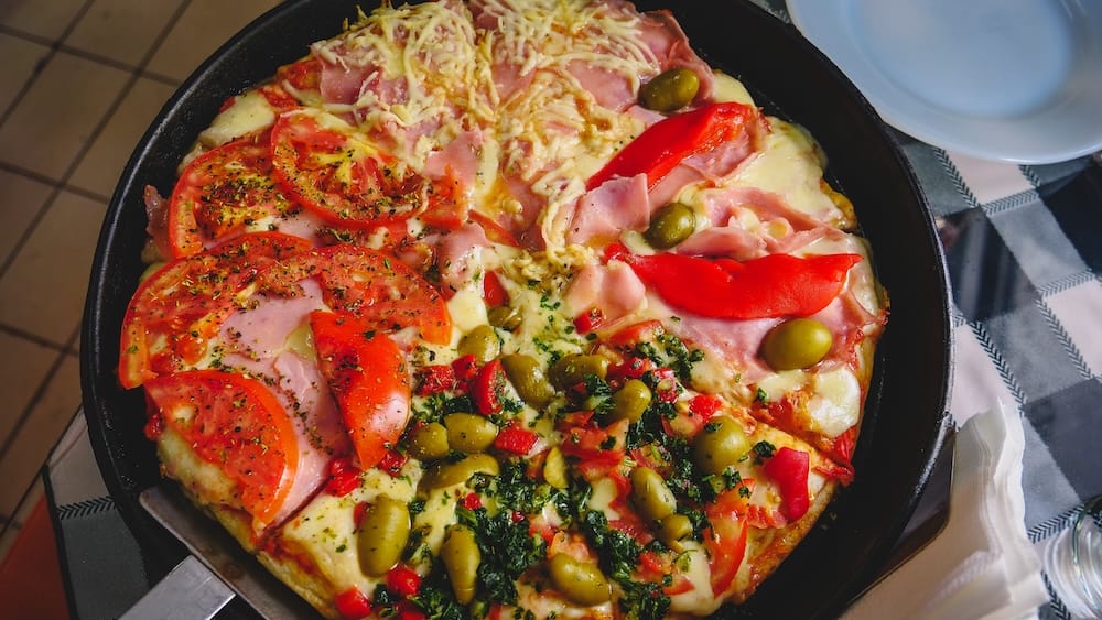 Quattro Stagioni pizza in Trelew Patagonia with ham olives tomato and peppers on thick crust in Argentina Quattro Stagioni pizza in Trelew Patagonia Argentina with ham, sliced tomatoes, olives, roasted red peppers, herbs, and melted cheese on a thick crust served in a casual inland pizzeria setting.