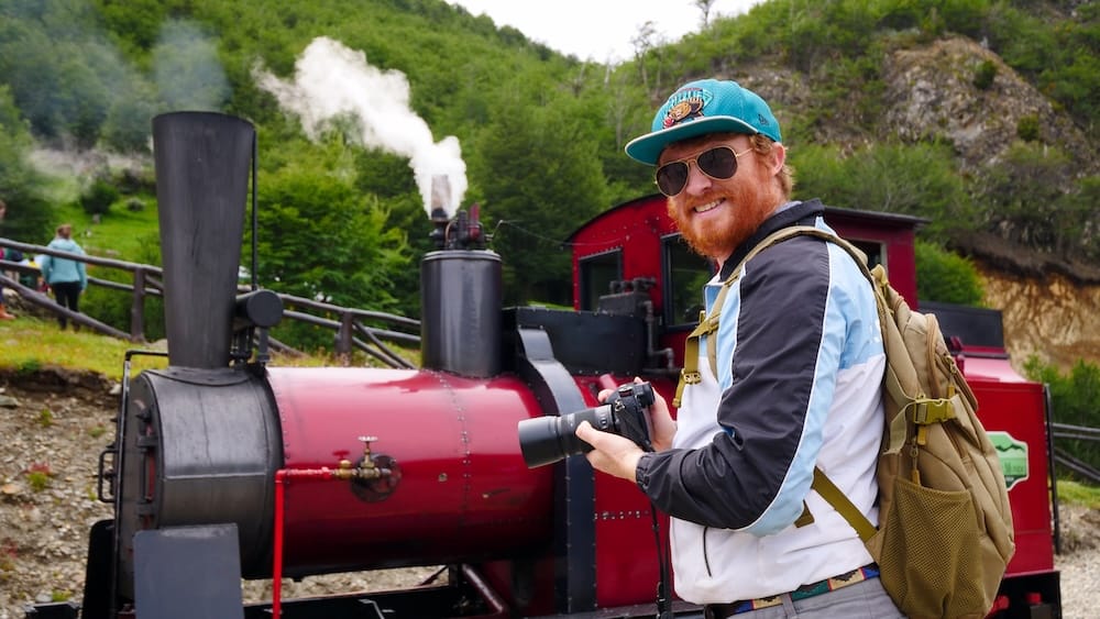 Samuel Jeffery holding a camera beside a red steam locomotive on the End of the World Train near Ushuaia, Tierra del Fuego, in Argentine Patagonia, with green hills and rising steam behind him.