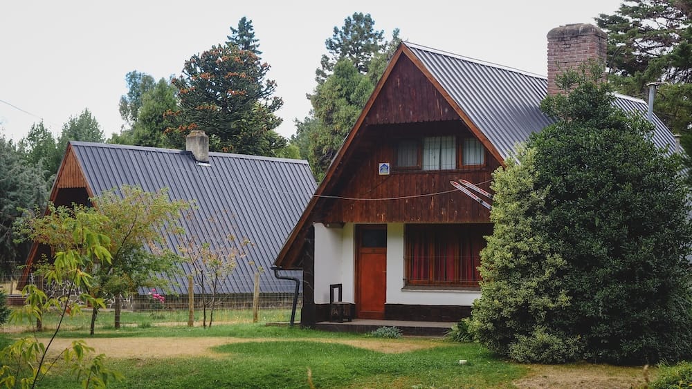 A-frame wooden cottages with steep metal roofs, chimneys and garden space in Trevelin, Chubut, Patagonia, Argentina, surrounded by trees and low mountain-town vegetation.