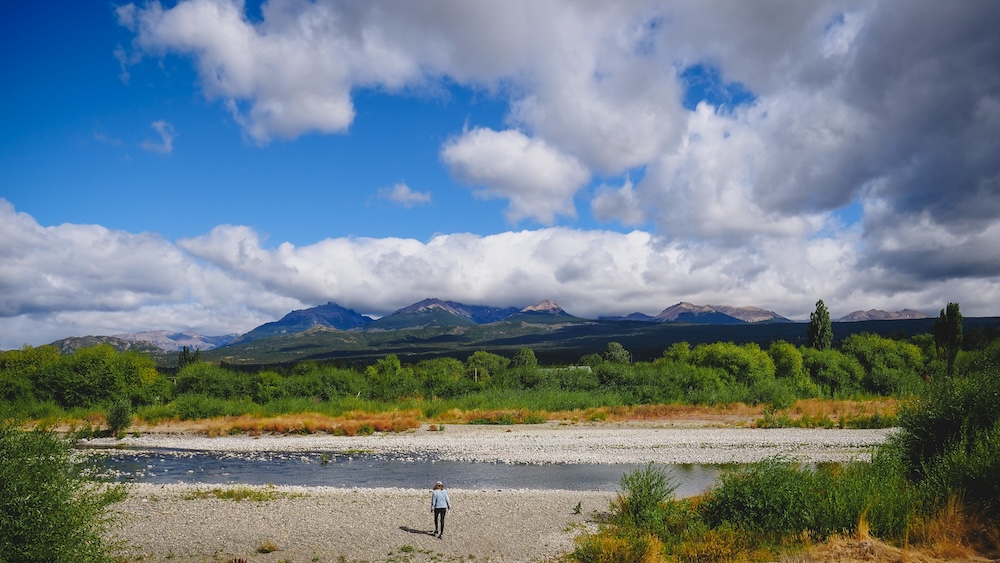 Trevelin Andean landscape in Chubut Patagonia Argentina with Audrey Bergner standing beside a wide riverbed surrounded by green valley fields and snow-dusted mountains, illustrating the lush alpine scenery of Welsh Patagonia near the town founded by Welsh settlers.