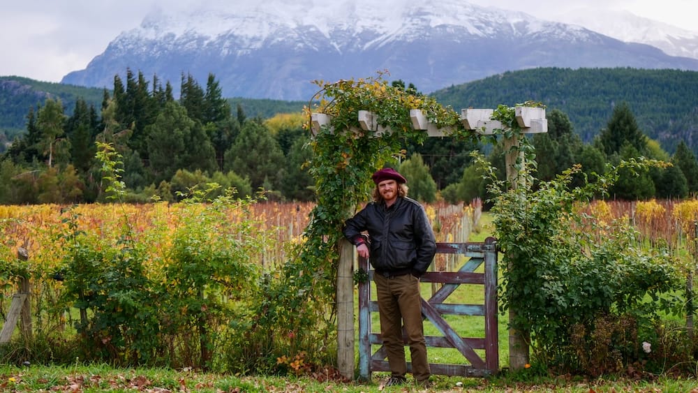 Autumn Vineyard in Trevelin, Chubut, Patagonia, Beneath the Andes with Samuel Jeffery Samuel Jeffery stands beside autumn vines in Trevelin, Chubut, Patagonia, Argentina, framed by a wooden garden gate with forested hills and snow-covered Andean peaks rising behind him.