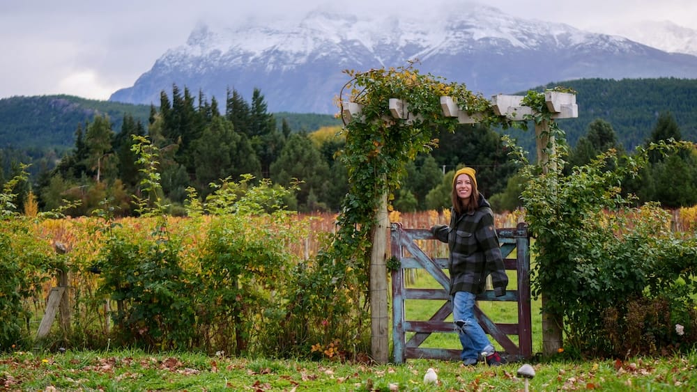Audrey Bergner at a Trevelin vineyard gate with snow-capped mountains in Chubut, Patagonia, Argentina Audrey Bergner stands by a wooden gate in a vineyard in Trevelin, Chubut, Patagonia, Argentina, with autumn vines, green fields and dramatic snow-capped mountains rising behind her under a cloudy sky.