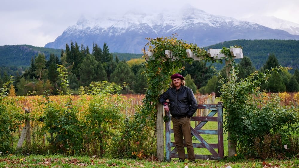 Trevelin Patagonia vineyard with Samuel Jeffery and Andes mountain backdrop Samuel Jeffery standing at a vineyard gate in Trevelin Patagonia Argentina with rows of vines, forest, and snow-capped Andes mountains in the background