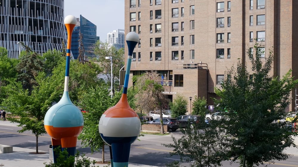 Dos de las esculturas cinéticas altas y coloridas "TRIO" afuera de la Biblioteca Central de Calgary en Calgary, Alberta, Canadá, que se asemejan a pájaros bebedores de gran tamaño, con edificios de gran altura y residenciales del centro al fondo.
