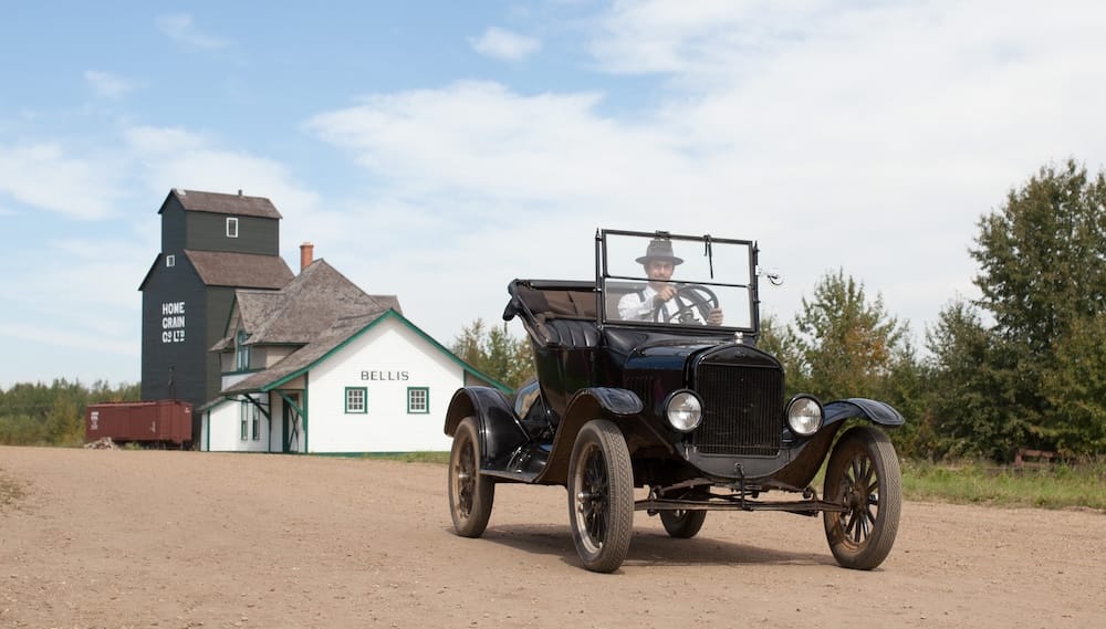 A historic black automobile drives past a grain elevator and heritage buildings at the Ukrainian Cultural Heritage Village in Alberta, Canada, with an interpreter behind the wheel.