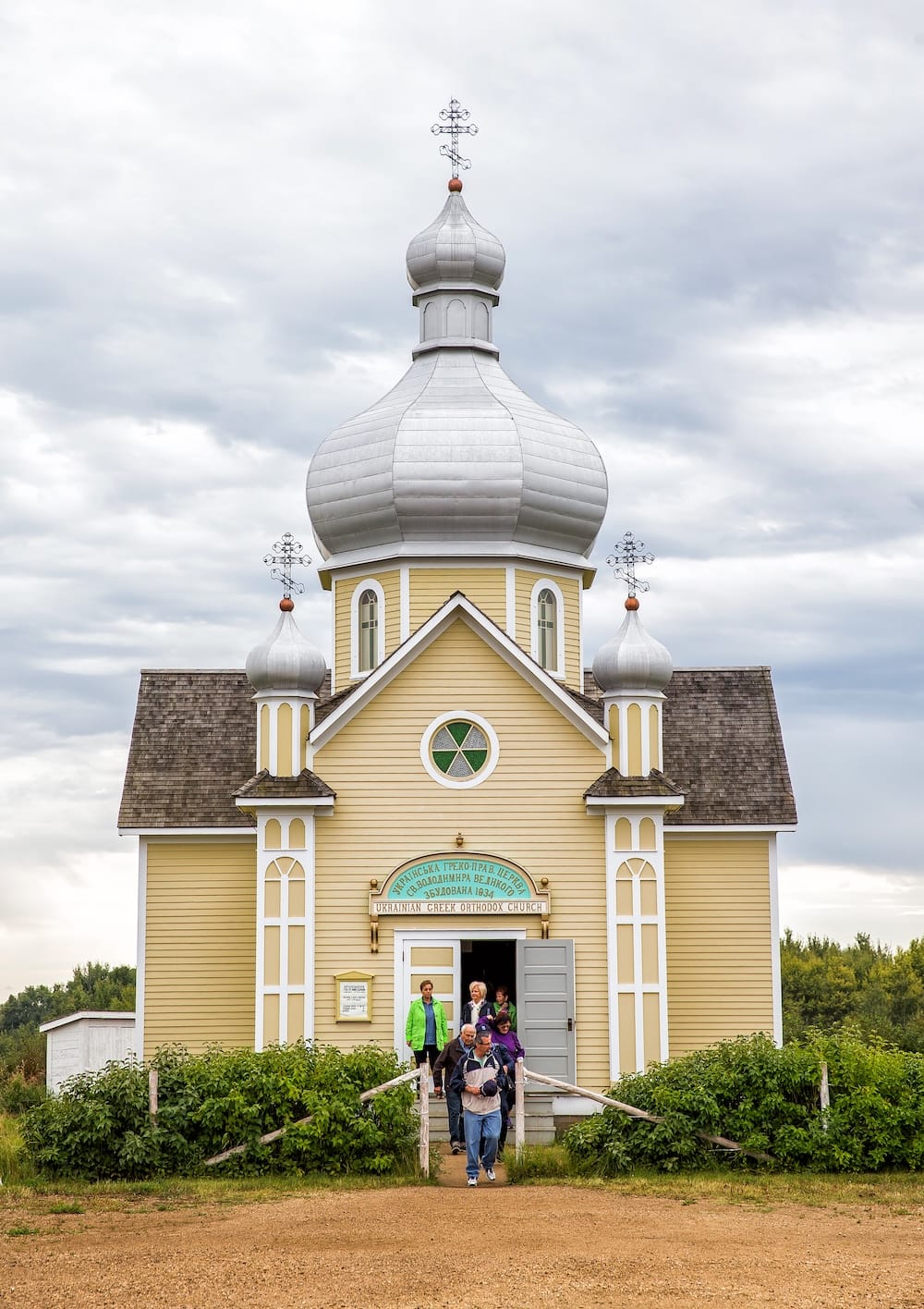 A yellow Ukrainian Greek Orthodox church with silver onion domes stands at the Ukrainian Cultural Heritage Village in Alberta, Canada, as visitors walk out beneath an overcast sky.