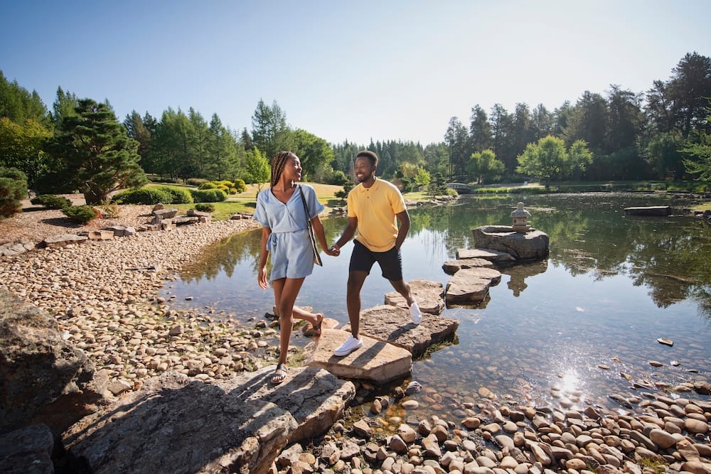 A couple walks across large stepping stones beside a landscaped pond at the University of Alberta Botanic Garden near Edmonton, Alberta, Canada, surrounded by trees, rocks and manicured gardens.