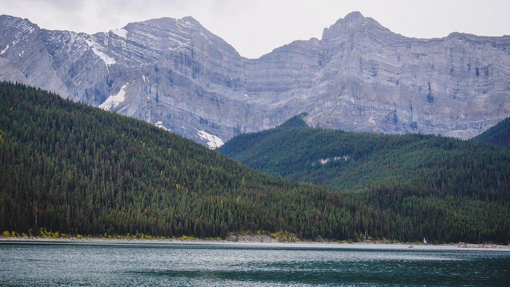 Upper Kananaskis Lake and surrounding peaks, Peter Lougheed Provincial Park, Alberta Snow-dusted rock peaks rise above dense forested slopes surrounding Upper Kananaskis Lake in Peter Lougheed Provincial Park, Alberta, with a small boat visible near the far shore.