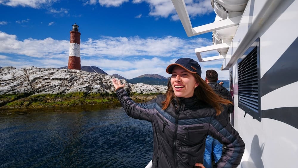 Ushuaia Beagle Channel boat tour with Audrey Bergner gesturing toward Les Éclaireurs Lighthouse, rocky islands, blue water, and mountains during Project 23 fieldwork in southern Tierra del Fuego, Argentina.