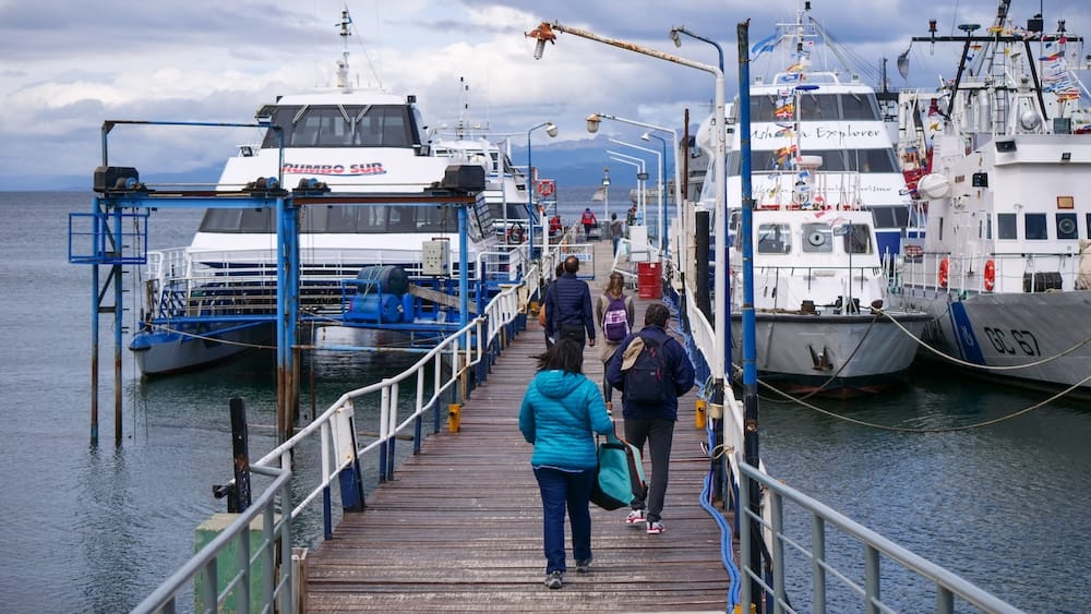 Travelers walk along the wooden maritime pier in Ushuaia, Tierra del Fuego, Patagonia, Argentina, toward catamarans and excursion boats preparing for Beagle Channel cruises under an overcast sky.