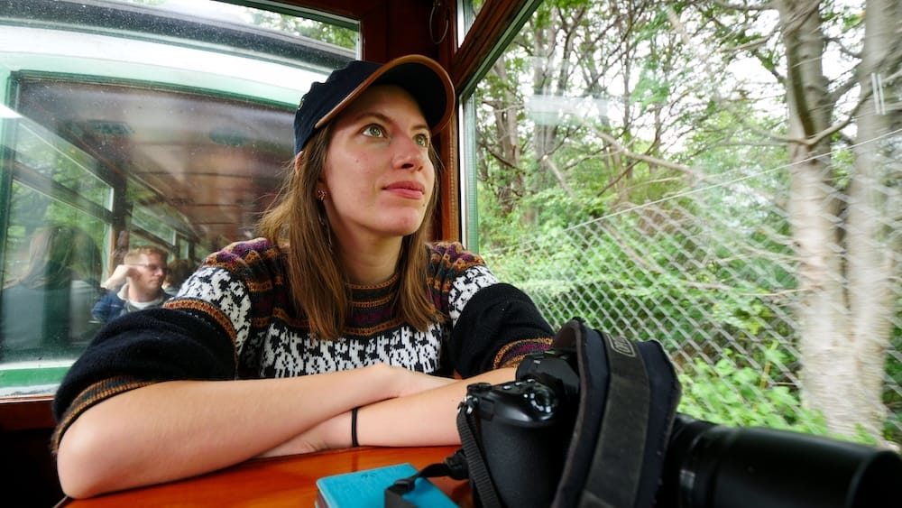Audrey Bergner rests her arms on a wooden table beside a camera, looking out the window of the End of the World Train in Tierra del Fuego National Park near Ushuaia, Argentina.