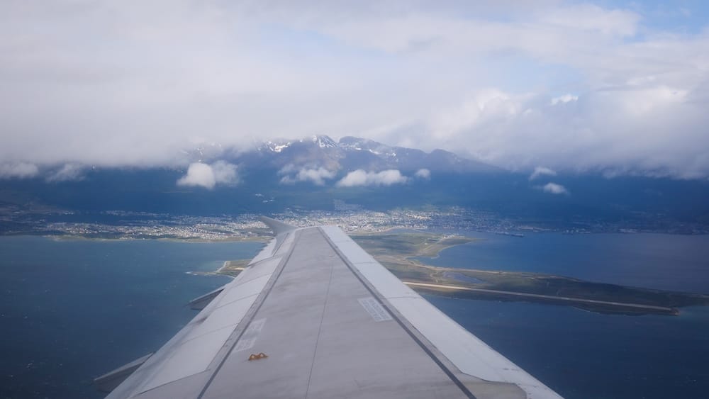Aerial Arrival View of Ushuaia, Tierra del Fuego, Patagonia, Argentina A view from an airplane wing descending toward Ushuaia, Tierra del Fuego, Argentina, showing the runway peninsula, the Beagle Channel, and cloud-wrapped snow-capped mountains beyond the town.