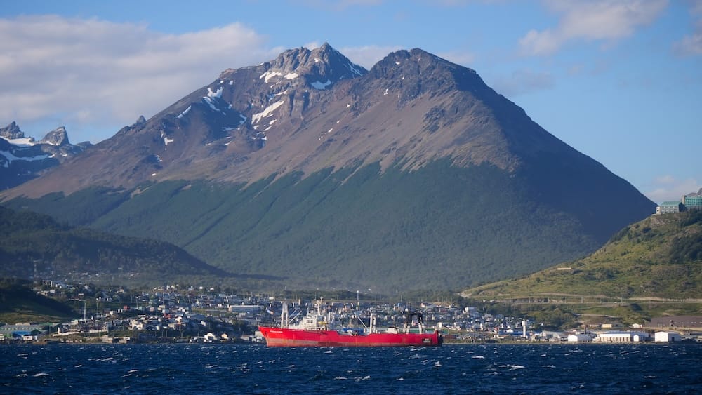 Un buque de carga rojo navega frente a Ushuaia, Tierra del Fuego, Patagonia, Argentina, con la ciudad extendida sobre la costa y montañas empinadas con restos de nieve detrás, bajo un cielo suave.