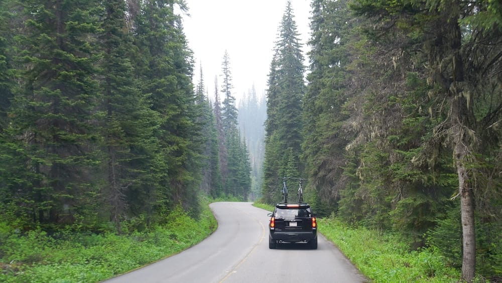 Una SUV oscura con bicicletas montadas en el techo maneja por una ruta sinuosa de montaña y bosque en el Parque Nacional Banff, Alberta, Canadá.