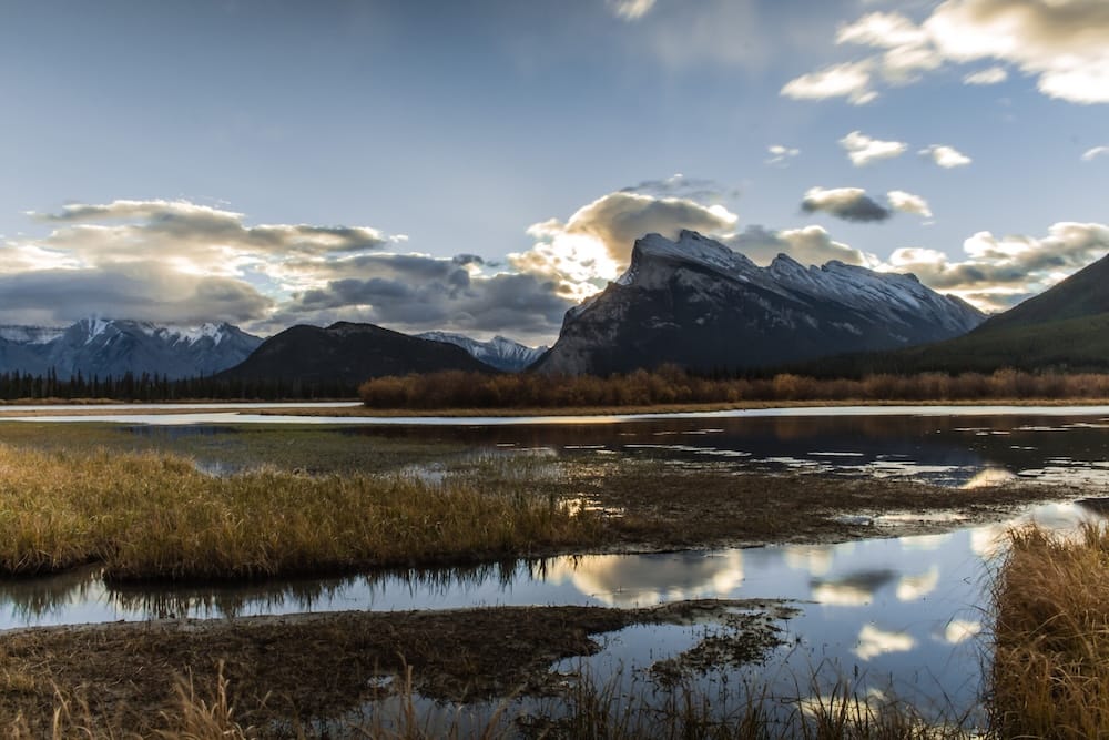 Mount Rundle rises above wetland grasses and still water at Vermilion Lakes, its rugged peak reflected in the surface under a cloudy evening sky near Banff, Alberta, Canada.