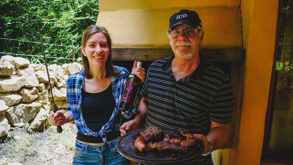 Villa Berna Argentine asado in Córdoba with Audrey Bergner and Daniel Bergner holding wine, grilling tools, and a platter of barbecued meat beside an outdoor parrilla, documenting local food culture and family fieldwork for Project 23.