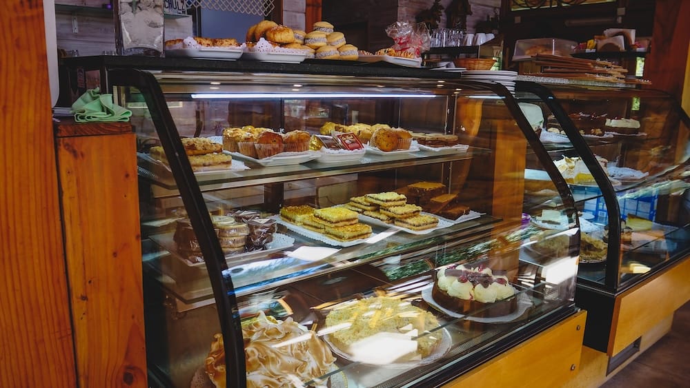 Patagonian Bakery Display in Villa La Angostura with Facturas Cakes and Assorted Pastries Glass display case filled with assorted Patagonian pastries and cakes in a Villa La Angostura bakery, capturing the overwhelming point-and-pick moment where travelers navigate facturas, slices, and sweets as practical fuel.