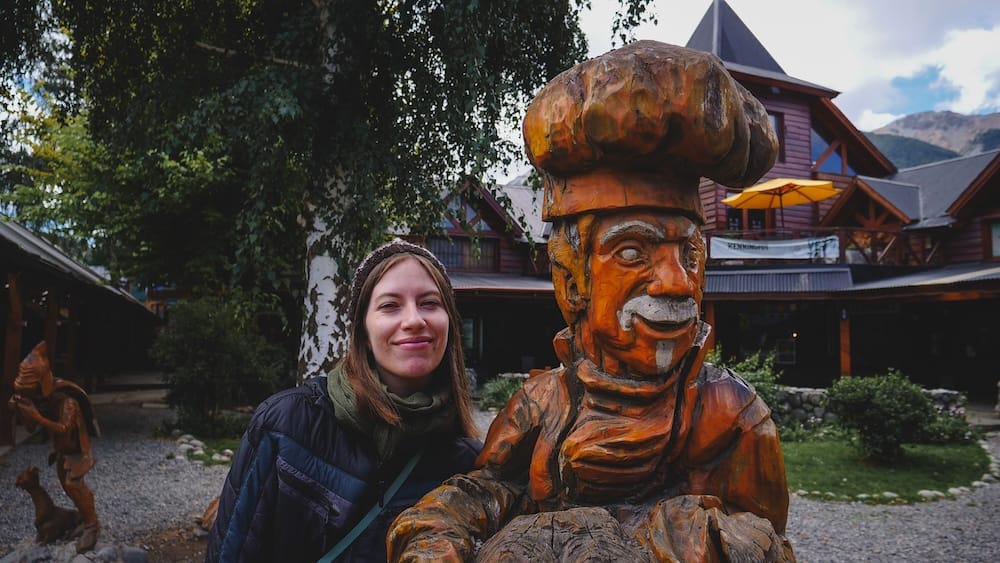 Audrey Bergner smiles beside a hand-carved wooden baker statue outside a chalet-style building in Villa La Angostura, Neuquén, Argentina, in the Patagonian Lake District.