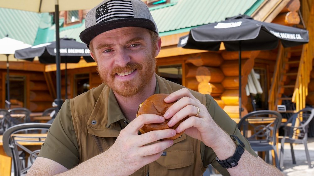 Nomadic Samuel eating a wagyu smashed burger at Bear Bistro in Island Lake Lodge near Fernie, BC Nomadic Samuel smiling while holding a wagyu smashed burger on the outdoor patio at Bear Bistro in Island Lake Lodge near Fernie, British Columbia, capturing one of the most memorable and indulgent meals he enjoyed during his Fernie food-focused mountain getaway.