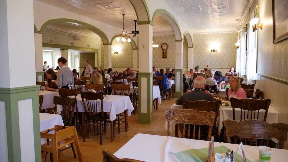The Wainwright Hotel dining room at Heritage Park in Calgary, Alberta, Canada, showing white-clothed tables, green-trimmed archways, and staff serving guests under a pressed-tin ceiling.