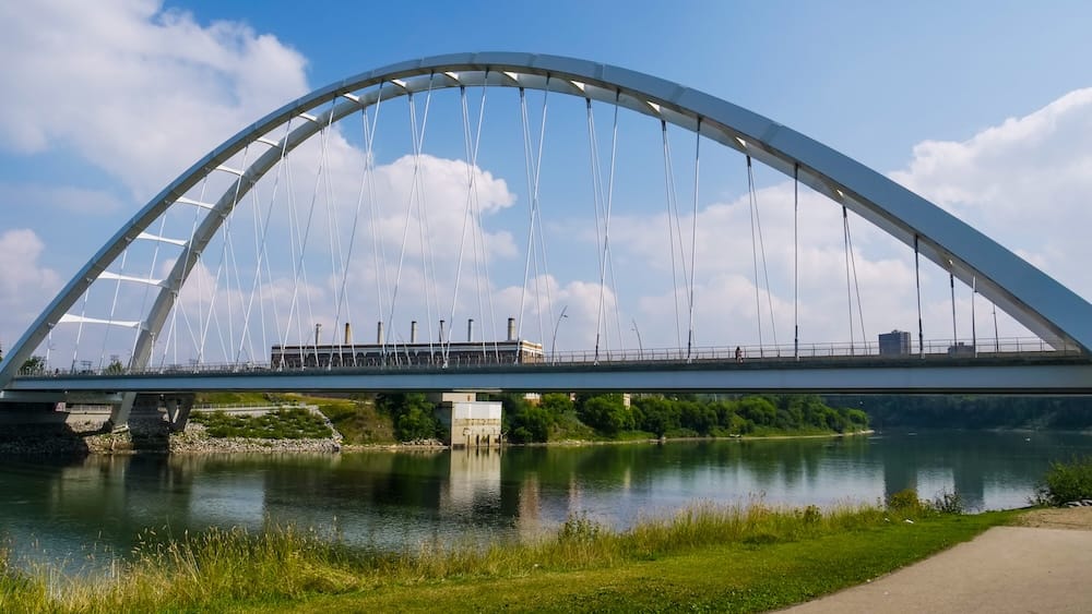 Walterdale Bridge sobre el North Saskatchewan River, red de senderos del valle del río en Edmonton, Alberta, Canadá Un puente blanco de arco atirantado, probablemente el Walterdale Bridge, cruza el North Saskatchewan River junto a la red de senderos del valle del río en Edmonton, con un camino pavimentado y el horizonte del centro de la ciudad de fondo, Alberta, Canadá