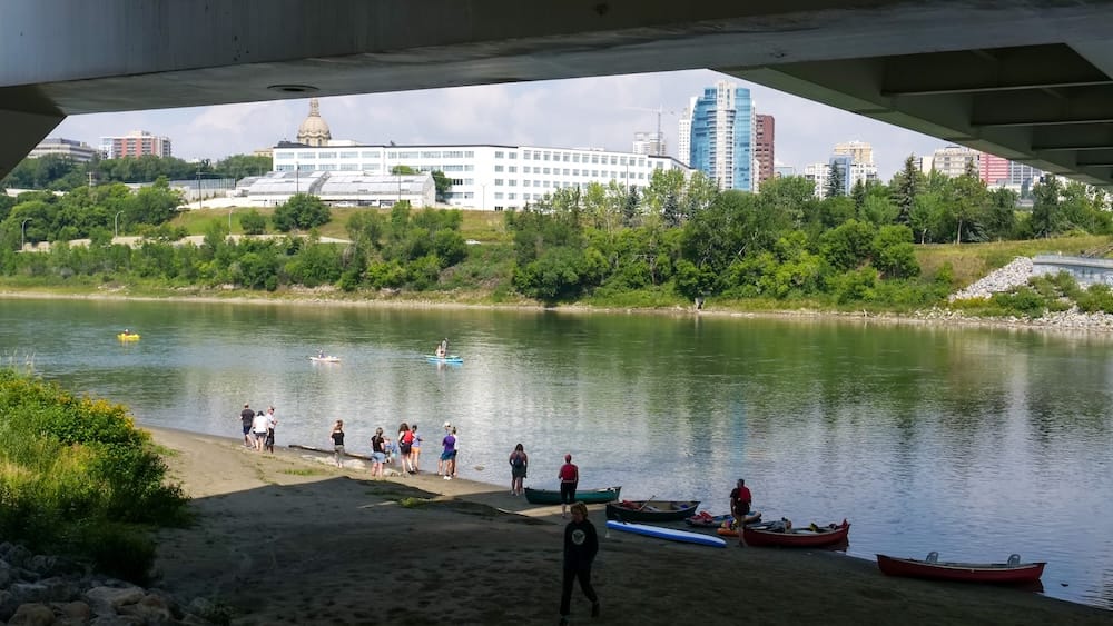 View from beneath Walterdale Bridge in Edmonton, Alberta, Canada, with canoes and paddleboarders gathered along the North Saskatchewan River and downtown buildings rising across the wooded bank.