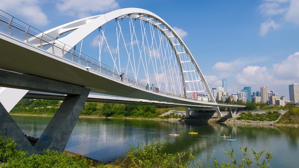Walterdale Bridge spanning the North Saskatchewan River in Edmonton, Alberta, Canada, with pedestrians crossing the white arch bridge and kayakers paddling below near the downtown skyline.