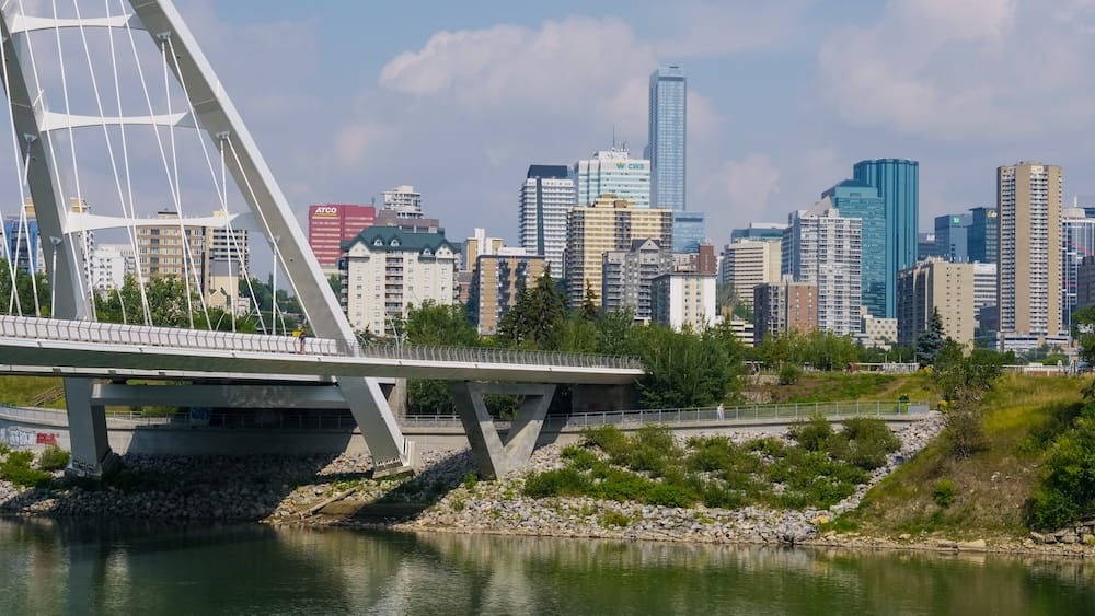 View across the North Saskatchewan River Valley in Edmonton, Alberta, Canada, with the white Walterdale Bridge in the foreground and the modern downtown skyline rising beyond the green riverbank.
