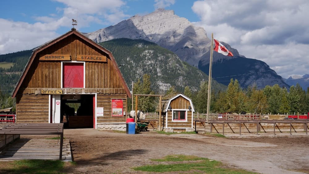 Warner Stables en Banff, Alberta, Canadá, con bandera canadiense y montañas de fondo antes del paseo en carreta Warner Stables en Banff, Alberta, Canadá, con su oficina rústica de madera, zona cercada para caballos, bandera canadiense y una gran montaña de las Rocosas bajo un cielo parcialmente nublado.