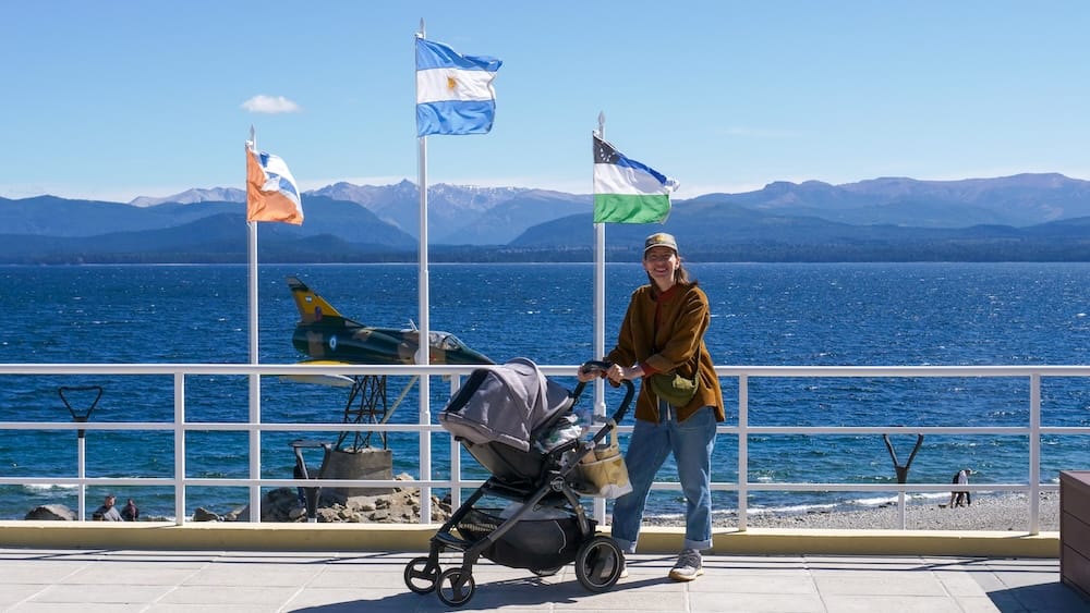 Audrey Bergner pushes toddler Aurelia Jeffery in a stroller along the waterfront promenade in San Carlos de Bariloche, Río Negro, Patagonia, Argentina, with lake, mountains, flags and an aircraft monument behind them.