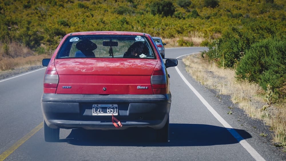 Weathered Red Sedan on a Winding Patagonia Road Near Cholila and the Deceptive Travel Times of Argentine Distances Weathered red sedan driving a winding paved road in Patagonia near Cholila, illustrating how even ordinary highway stretches toward events like Fiesta Nacional del Asado can take longer than maps suggest because of curves, traffic and road conditions.