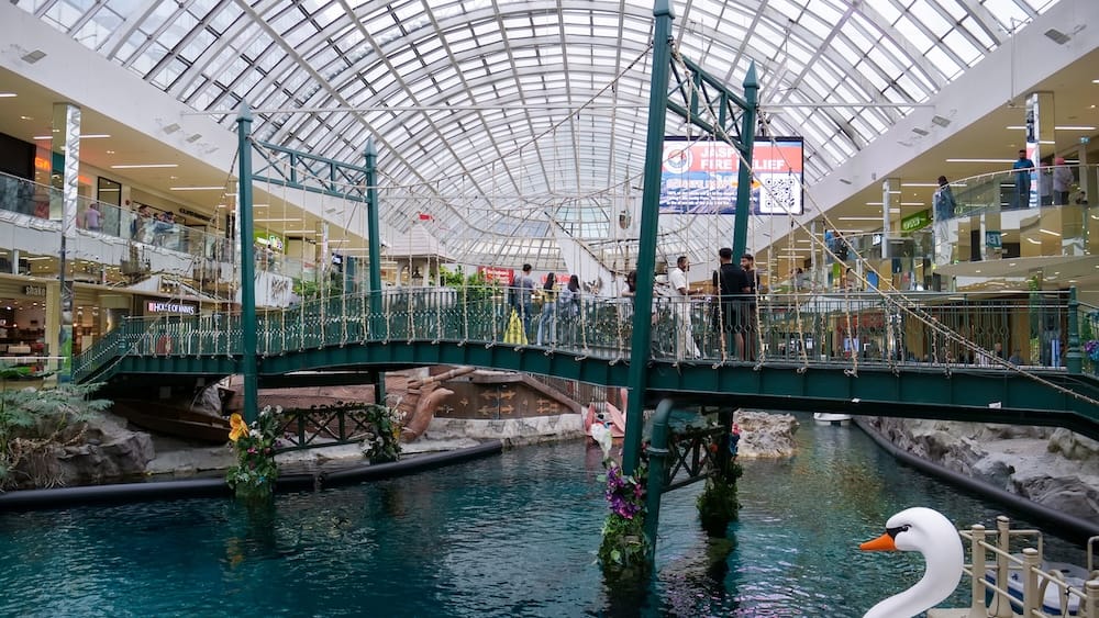 Indoor lagoon and decorative suspension bridge inside West Edmonton Mall in Edmonton, Alberta, Canada, with shoppers on two levels beneath the curved glass roof.