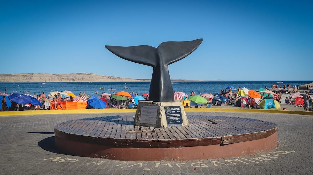 A whale tail monument overlooks a crowded summer beach in Puerto Pirámides on Península Valdés, Chubut, Patagonia, Argentina, with colourful umbrellas, swimmers and calm blue water beyond.