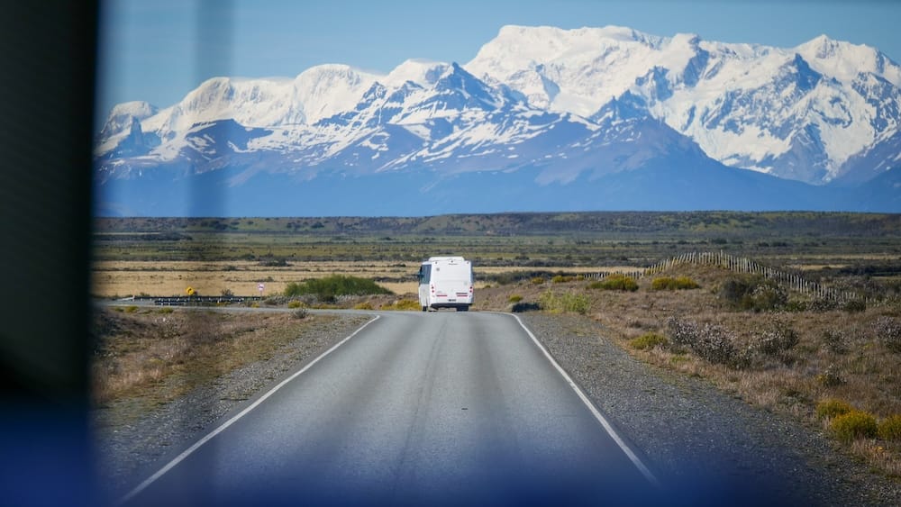 White Bus on the Highway Near El Calafate, Santa Cruz, Patagonia, Argentina, Beneath Snowy Mountains A white bus drives along a lonely highway near El Calafate in Santa Cruz, Patagonia, Argentina, crossing the open steppe toward a dramatic wall of snow-covered mountains seen from inside another long-distance coach.